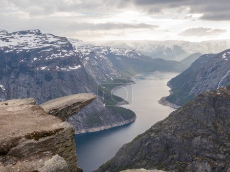 Téléchargez les photos : Famous rock formation, Trolltunga with a view from the above on Ringedalsvatnet lake, Norway. Rock hanging. Slopes of the mountains are partially covered with snow. The water of the lake is navy blue. - en image libre de droit