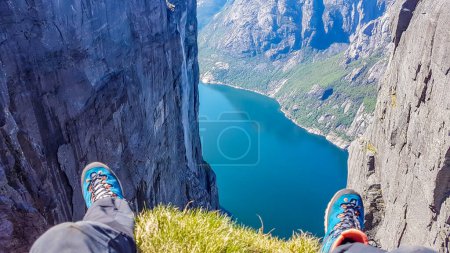 Foto de Dos pies con botas de senderismo en ellos colgando de una roca escarpada con vistas a la impresionante Lysefjorden brillante con muchos tonos de azul y verde. Pendientes empinadas de la montaña, cascada corriendo hacia abajo - Imagen libre de derechos