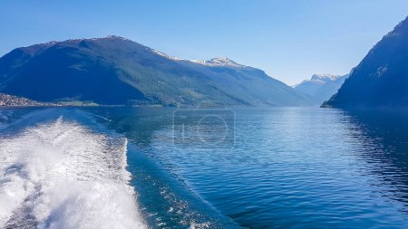 Téléchargez les photos : A view on the Songefjorden fjord from the water level. The motor of the ship makes the water wavy and foamy. Tall, lush green mountains surrounding the fjord. Clear blue sky. - en image libre de droit