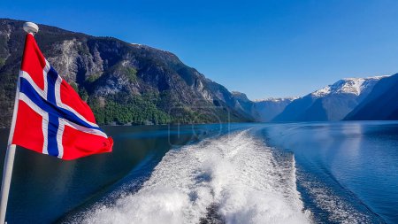 Téléchargez les photos : Norwegian flag hanging on  the railing of the ship and waving above the water.The motor of the ship makes the water wavy and foamy. Tall, lush green mountains surrounding the fjord. Clear blue sky. - en image libre de droit
