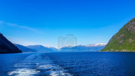 Téléchargez les photos : Une vue sur le fjord de Songefjorden depuis le niveau d'eau. Le moteur du navire rend l'eau ondulée et mousseuse. De hautes montagnes verdoyantes entourant le fjord. Ciel bleu clair. - en image libre de droit