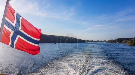 Téléchargez les photos : Drapeau norvégien accroché sur la rambarde du navire et agitant au-dessus de l'eau.Le moteur du navire rend l'eau ondulée et mousseuse. De hautes montagnes verdoyantes entourant le fjord. Ciel bleu clair. - en image libre de droit