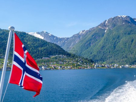 Téléchargez les photos : Drapeau norvégien accroché sur la rambarde du navire et agitant au-dessus de l'eau.Le moteur du navire rend l'eau ondulée et mousseuse. De hautes montagnes verdoyantes entourant le fjord. Ciel bleu clair. - en image libre de droit