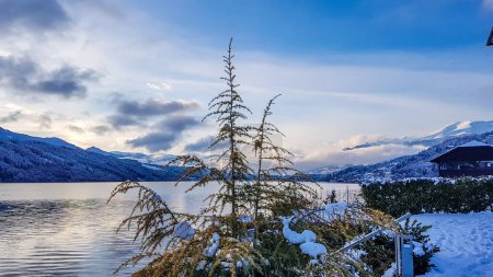 Photo for A beautiful view on a Millstaetter lake in Austria. The lake is surrounded by Alps. Mountains are covered with snow. Soft colors of the sunset. Blue sky. In the middle there is a tree. - Royalty Free Image