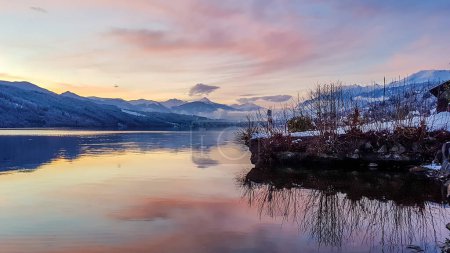 Photo for Beautiful view on Millstaetter lake in Austria. The lake is surrounded by Alps. Mountains are covered with snow. The sky is exploding with pink and orange. Stunning sunset. Shore overgrown with bushes - Royalty Free Image