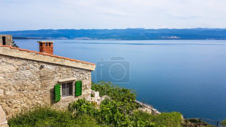 Téléchargez les photos : Une maison en pierres avec volets verts située sur une colline avec une vue magnifique sur la mer. Chaque coin de la maison est recouvert de petites vignes et de buissons. Surface calme de la mer. Gemme cachée - en image libre de droit