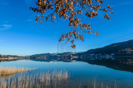 Téléchargez les photos : Une vue sur un lac et les Alpes à l'arrière. La surface calme du lac reflète les montagnes, les rayons de soleil et les nuages. Petite ville située au bord du lac. Eglise avec clocher au milieu - en image libre de droit