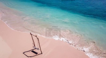 Foto de A swing placed on the seashore of Pink Beach, Lombok, Indonesia. The swing has very simple wood construction. Waves gently wash the pillars of it. In the back there are few boats. Drone capture. - Imagen libre de derechos