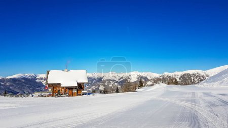 A chalet in Bad Kleinkirchheim, Austria. The little cottage is located next to a ski slope, surrounded by thick snow. The slope is perfectly gravelled. There are snow caped mountains in the back.