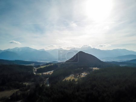 Téléchargez les photos : Vue imprenable sur Kathreinkogel à Schiefling am See, Autriche. La colline a une forme pyramidale parfaite. Il est envahi par la forêt. Il y a de hautes Alpes à l'arrière. Petit village sous la colline. - en image libre de droit