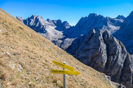 Foto de A pathway mark on the trail to the Grosse Gamswiesenspitze in Lienz Dolomites, Austria. Sharp peaks in the back. Massive Alpine mountain. Slopes have golden color. Making a decision which way to go - Imagen libre de derechos