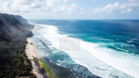 Eine Aufnahme von Nyang Nyang Beach, Bali, Indonesien. Die Wellen rauschen ans Ufer und lassen das Wasser sprudeln. Der Strand ist mit Grünalgen bedeckt, weiter unten ist er sandig. Hohe Klippen an der Seite