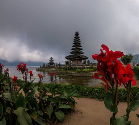 Foto de Vista en la parte delantera del edificio principal del Templo Ulun Danu, Bali, Indonesia, a través de flores rojas. Hermoso templo de agua hindú. Lugar sagrado. Descubriendo nuevas culturas. El templo está rodeado de agua - Imagen libre de derechos