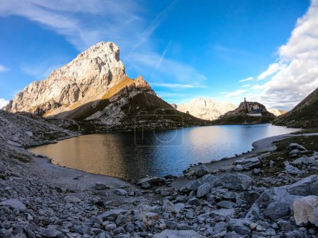 Photo for Panoramic shot of a reflection of an Alpine mountain in Wolayer Lake Austria. Completely still surface of the lake. Mountain is catching the first sunbeams. Alpine cottage on the side. High altitude - Royalty Free Image