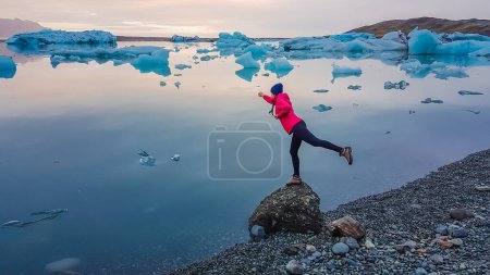 A girl wearing pink jacket stands at the rock by the glacier lagoon, forms a flying superhero pose. Empowered woman. Blue icebergs slowly drifting towards the sea. Effect of global warming.