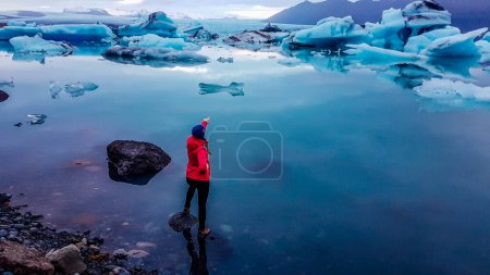 Foto de Una chica con chaqueta rosa de pie sobre la roca en un agua poco profunda. Ella está señalando a los bergs de hielo a la deriva en la laguna glaciar. El glaciar tiene muchos tonos de azul. Calentamiento global que causa el derretimiento de glaciares - Imagen libre de derechos