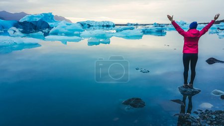 Foto de Una chica con chaqueta rosa de pie sobre la roca en un agua poco profunda. Ella extiende sus brazos hacia arriba y ancho. Laguna glaciar llena de helados a la deriva. Calentamiento global que causa el derretimiento de glaciares. Tonos fríos. - Imagen libre de derechos