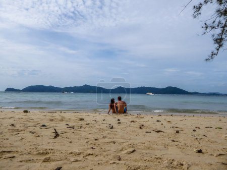 Foto de Una pareja sentada a la orilla de una playa de arena. Llevan trajes de baño. Otras islas visibles en la espalda. Clima claro y agradable. Relajación en la orilla. Algunas ramas de árboles visibles en el lado. - Imagen libre de derechos