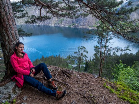 Photo for A girl wearing a pink pullover leans against a tree and takes he rest. The tree branches are spreading around. Marvelous view on calms water of a fjord from above. Hiking in a fjord. - Royalty Free Image