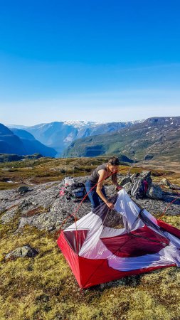 Téléchargez les photos : A girl in hiking outfit struggles to out the tent up together. She is camping in the wilderness. The backdrop is Eidfjord and tall glacier. Adventurous solo traveling girl. Bright and sunny day - en image libre de droit