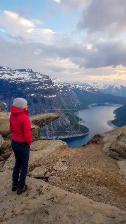 Foto de A girl wearing pink jacket stands next to the hanging rock formation Trolltunga with a view on Ringedalsvatnet lake, Norway at the sunrise. Slopes are partially covered with snow Freedom and happiness - Imagen libre de derechos