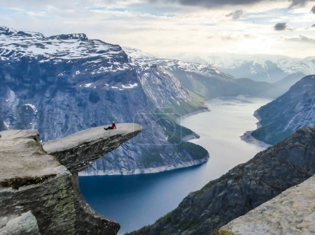 Foto de A girl wearing pink jacket stands at the hanging rock formation, Trolltunga with a view on Ringedalsvatnet lake, Norway. Slopes of the mountains are partially covered with snow. Freedom and happiness - Imagen libre de derechos