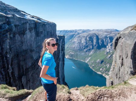 Téléchargez les photos : Une fille portant un T-shirt bleu lève les yeux, sur les falaises environnantes. Elle profite de la vue sur le fjord devant elle. Superbe Lysefjorden brille avec de nombreuses nuances de bleu et de vert. La fille est heureuse après la randonnée - en image libre de droit