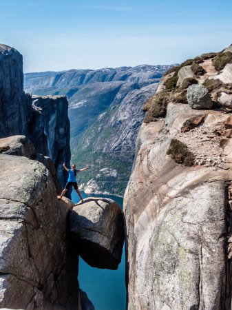 Foto de Una chica con una camiseta azul de pie con una pierna colgando de una roca, Kjerag, Noruega. La roca en forma de bola está colgando entre paredes de piedra con una caída libre de 1000m. Ella es concentrada y cautelosa. - Imagen libre de derechos