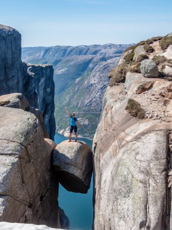 Foto de Una chica con una camiseta azul de pie en la roca colgante, Kjerag, Noruega. La roca en forma de bola está colgando entre paredes de piedra con una caída libre de 1000m. Felicidad y libertad después de pisar la roca - Imagen libre de derechos