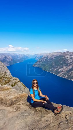 Photo for A girl wearing blue T-shirt sitting at the edge of a steep cliff of Preikestolen, with a view on Lysefjorden. Fjord goes far inland. Girl enjoys the view, feeling free and happy. Great accomplishment - Royalty Free Image