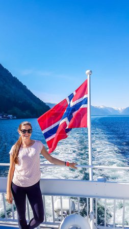 Téléchargez les photos : A young woman leaning on a rail of a ship with a Norwegian flag waving behind him, Songefjorden Norway. The motor of the ship makes the water wavy and foamy. Lush green mountains surrounding the fjord - en image libre de droit