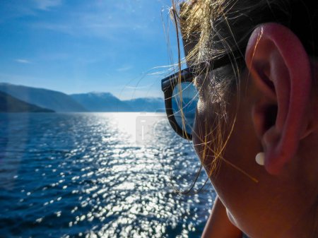 Téléchargez les photos : Un gros plan sur le visage de la jeune fille, prenant un bateau à travers les Songefjorden, Norvège. La fille admire le magnifique fjord. Le soleil se reflète dans l'eau. Des montagnes plus hautes couvertes de neige. Ciel clair. - en image libre de droit