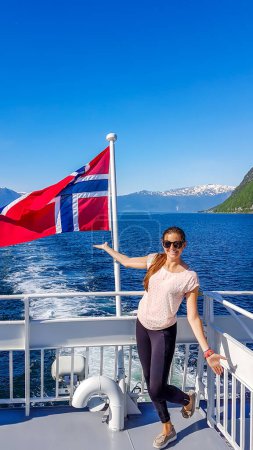 Téléchargez les photos : A young woman leaning on a rail of a ship with a Norwegian flag waving behind him, Songefjorden Norway. The motor of the ship makes the water wavy and foamy. Lush green mountains surrounding the fjord - en image libre de droit