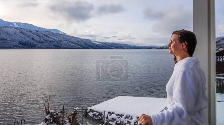 Photo for A girl in a white bathrobe standing on the balcony of a SPA in Austria in winter. She has wet hair. She is enjoying the view on Millstaetter lake and mountains in front of her. She id feeling relaxed - Royalty Free Image
