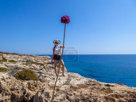Foto de A girl holding a gigantic flower in her hand. Changing the perspective, fun photography. Girl is wearing a hat and shorts. Behind there is an open sea. Barren landscape of Cape Greco, Cyprus. - Imagen libre de derechos