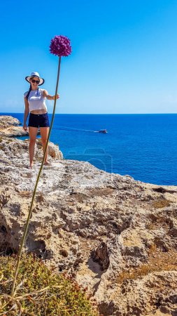 Foto de A girl holding a gigantic flower in her hand. Changing the perspective, fun photography. Girl is wearing a hat and shorts. Behind there is an open sea. Barren landscape of Cape Greco, Cyprus. - Imagen libre de derechos