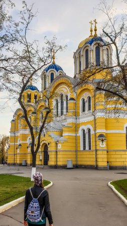 Téléchargez les photos : Une fille avec un sac à dos marchant vers la cathédrale St Volodymyr. La cathédrale est peinte en jaune, avec un toit en dôme bleu et doré. Jour couvert et froid. - en image libre de droit