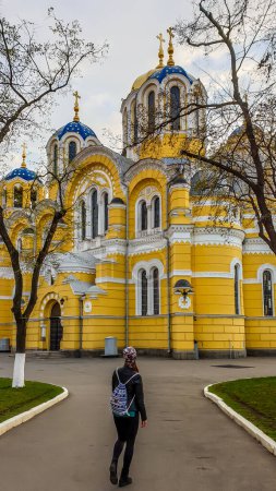 Téléchargez les photos : Une fille avec un sac à dos marchant vers la cathédrale St Volodymyr. La cathédrale est peinte en jaune, avec un toit en dôme bleu et doré. Jour couvert et froid. - en image libre de droit