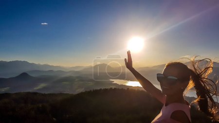 A girl in sunglasses trying to give a high five to the sun. The wind blows her hair in all the directions. There is a lake in the bottom of a valley. Girl is smiling and enjoying her time.