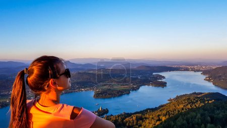 Téléchargez les photos : Une fille debout sur une terrasse d'observation et profitant de la vue imprenable sur la vallée et le lac Woerthersee pendant le coucher du soleil. Hills attrape les derniers rayons du soleil. Contemplation de la nature - en image libre de droit