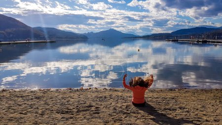 Téléchargez les photos : Girl wearing orange jumper, sitting at the sand beach by the Woertersee, Poertschach, Austria and flipping her hair high up. Sky reflects in the lake's surface. Alps surrounding the lake. Overcast. - en image libre de droit