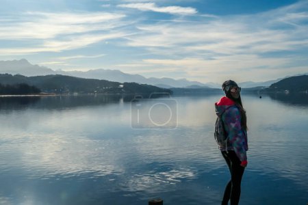 Téléchargez les photos : Girl wearing a jacket and beanie stands at the end of a promenade on the lake. Soft reflections of the clouds in the lake. Clear but cloudy day. High mountains in the back Calmness and relaxation - en image libre de droit