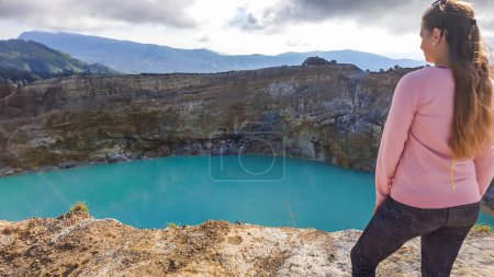 Foto de Una mujer parada en el borde del volcán y observando los lagos del cráter volcánico Kelimutu en Moni, Flores, Indonesia. La mujer está relajada y tranquila, disfrutando de la vista sobre el lago turquesa. Grande nublado. - Imagen libre de derechos