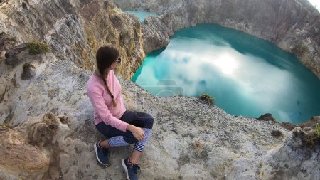 Foto de Mujer sentada en el borde del volcán y observando los lagos del cráter volcánico Kelimutu en Moni, Flores, Indonesia. La mujer está relajada y tranquila, disfrutando de la vista sobre el lago que brilla con muchos tonos de turquesa - Imagen libre de derechos