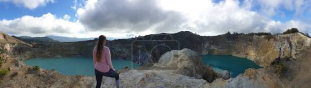 Foto de Una mujer parada en el borde del volcán y observando los lagos del cráter volcánico Kelimutu en Moni, Flores, Indonesia. La mujer está relajada y tranquila, disfrutando de la vista sobre el lago turquesa. Grande nublado. - Imagen libre de derechos