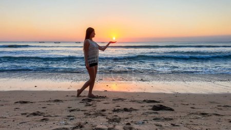 Foto de A girl holding a setting sun in her hand on Seminyak beach on Bali, Indonesia. The sun sets directly into the water. Calm sea washes gently the shore. The sunbeams reflecting on the sea surface. Fun - Imagen libre de derechos