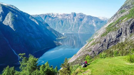 A couple standing on the meadow with a majestic view on Eidfjord from Kjeasen, Norway. Slopes of the mountains are overgrown with lush green grass. Water has dark blue color. Sunny and clear day