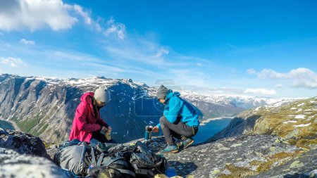 Foto de A couple having breakfast in the tall mountains with a view on a fjord-like lake. Girl is cutting the bread and the boy boils the water. Clear and sunny weather. Camping in the wilderness. - Imagen libre de derechos
