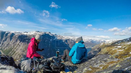 Foto de A couple having breakfast in the tall mountains with a view on a fjord-like lake. They are both relaxed and chilled. Clear and sunny weather. Camping in the wilderness in beautiful nature. - Imagen libre de derechos
