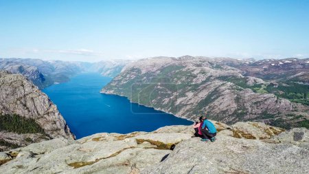 Téléchargez les photos : Couple assis sur un rocher et admirant une vue sur Lysefjorden. Prise de vue réelle, perspective supérieure sur le couple. Vue sans fin sur le fjord. Couple assis à l'écart des sentiers battus, liberté en randonnée. - en image libre de droit